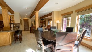 A spacious dining area with a large wooden table and chairs, a kitchen with wooden cabinets, and a view of the outdoors through large windows.