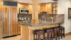 A kitchen with wooden cabinets, a central island with stools, a stainless steel refrigerator, and a microwave.