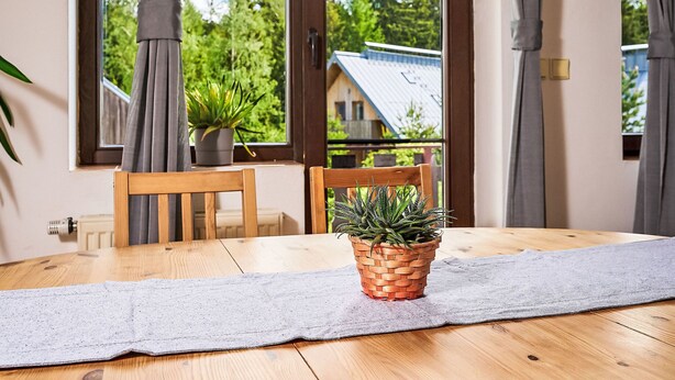 A dining area with a wooden table, two chairs, a woven plant pot, and a view of a house through large windows. - Alice II. by Interhome (Harrachov)