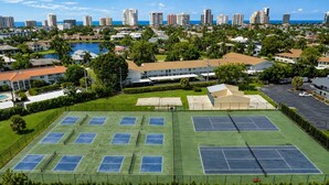 Un complexe de courts de tennis comprenant plusieurs terrains, entouré de bâtiments résidentiels et bordé par un plan d’eau.