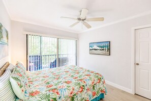 Une chambre avec un lit, un ventilateur de plafond, une porte-fenêtre coulissante donnant sur un balcon et un tableau encadré au mur.