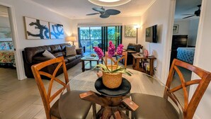 A modern living room with a dining area, featuring a ceiling fan, a large window, and a view of the outdoors.