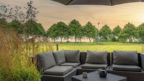 A wicker sofa with grey cushions and a table with a teapot, set under an umbrella, overlooking a grassy field with trees.