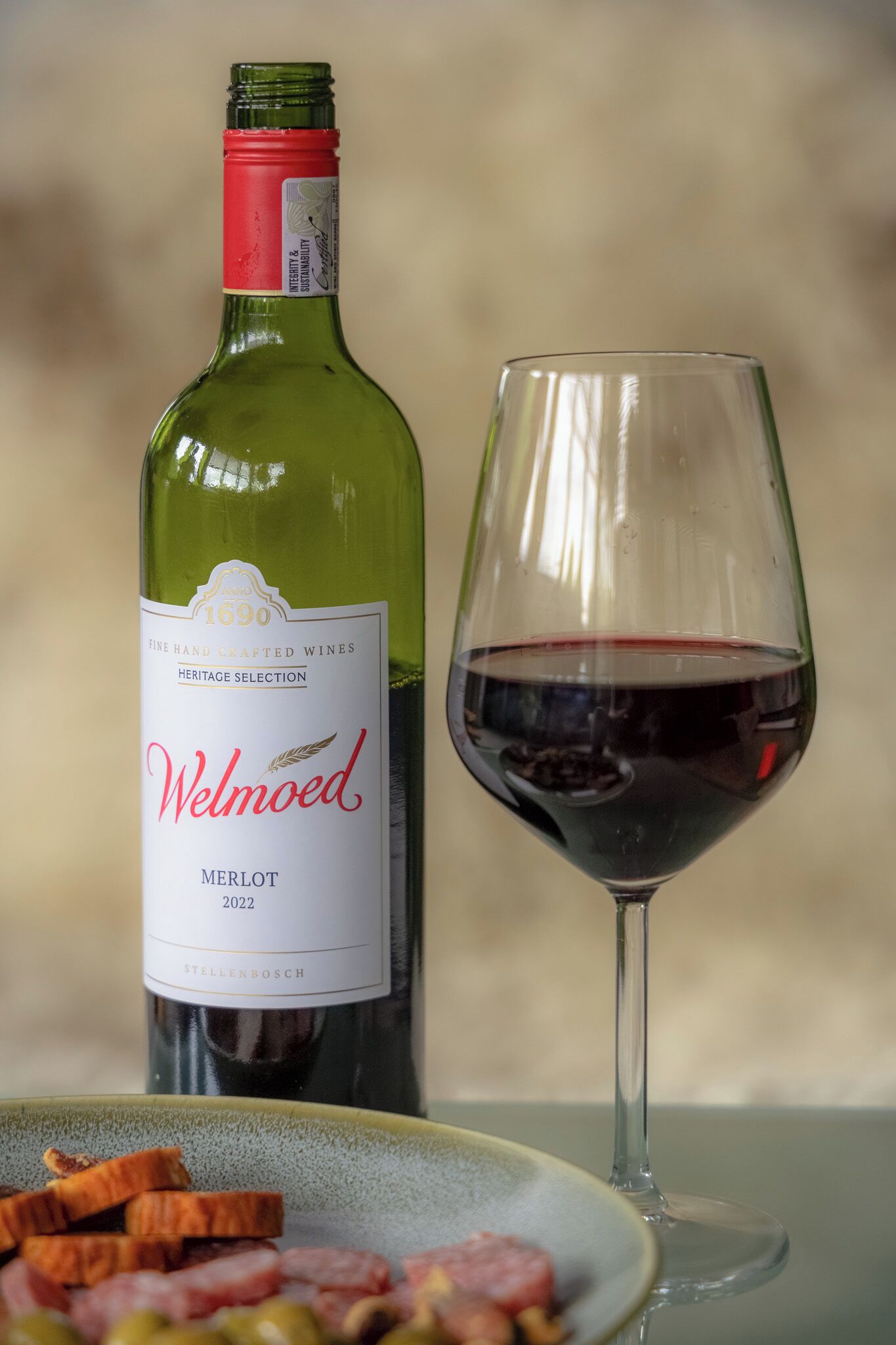 Una botella de vino tinto Welmoed Merlot 2022 junto a una copa llena de vino tinto, sobre una mesa con un plato de comida al frente.