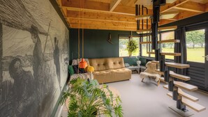 A modern living room with a high ceiling, wooden beams, a large mural, a sofa, a coffee table, a hanging plant, and a unique staircase.