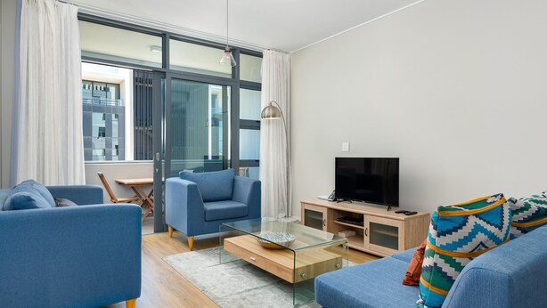 Ein modernes Wohnzimmer mit einem Glastisch, blauen Sofas und einem Flachbildfernseher auf einem Holzboden.