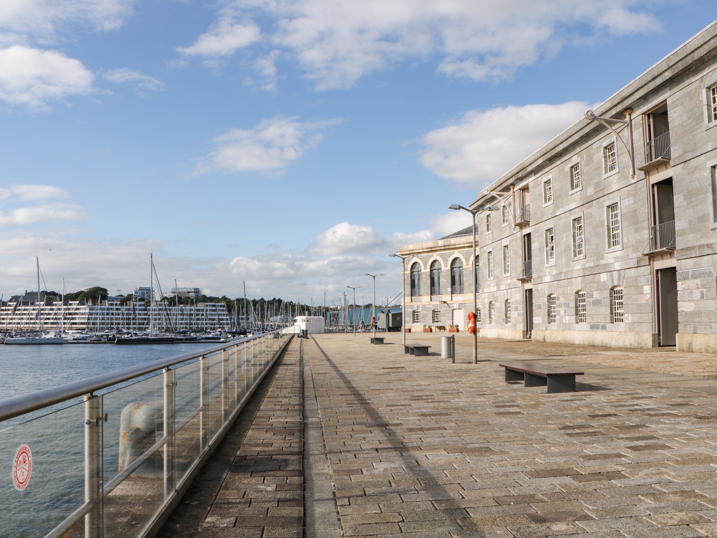 A waterfront promenade with a stone walkway, benches, and a historic building with arched windows.