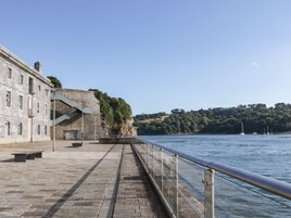 A waterfront promenade with a stone building, a metal staircase, and a railing overlooking a body of water with boats.