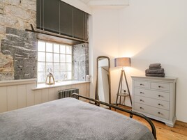A bedroom with a stone wall, a window with blinds, a bedside lamp, a dresser, and a bed with a textured blanket.