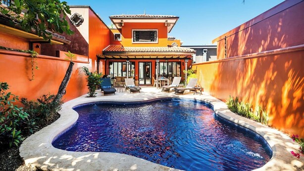 Ein Schwimmbecken mit Liegestühlen und einer Terrasse vor einem Gebäude mit orangefarbenen Wänden und einem Ziegeldach.