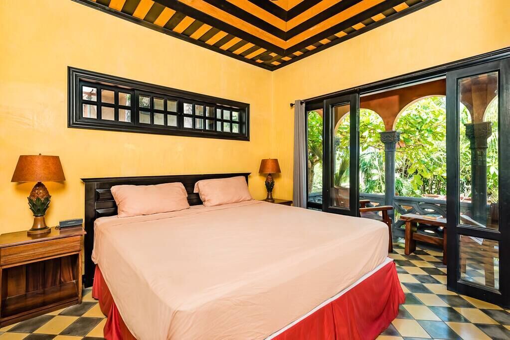 A hotel room with a large bed, two bedside tables with lamps, a window with a view of greenery, and a checkered floor.