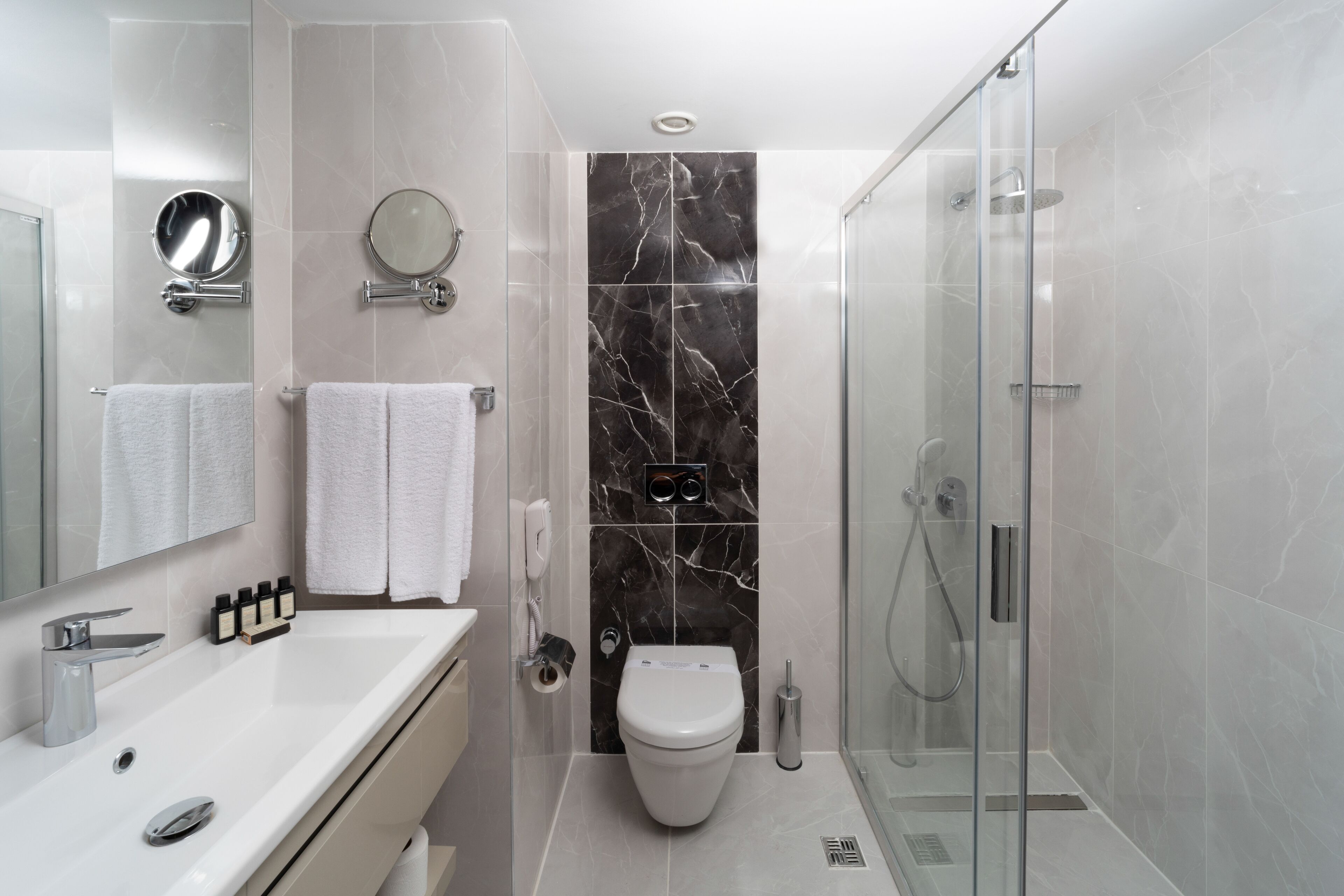 Et moderne badeværelse med en hvid vask, to spejle, et toilet og en bruseniche med glasvægge.