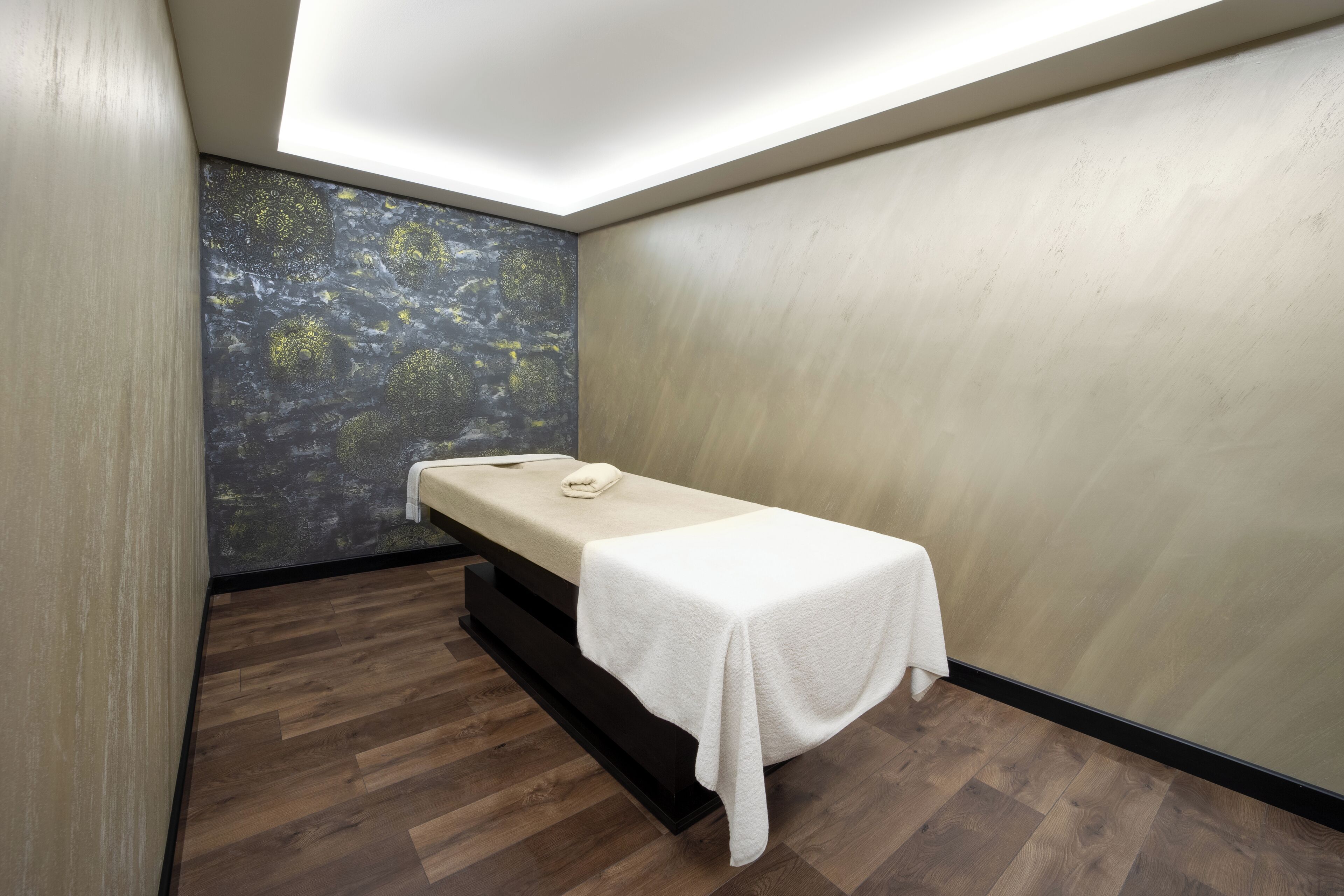 A treatment room with a wooden floor, a massage table, a wall with a nature-themed mural, and a ceiling light.