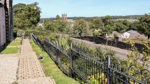 A pathway leading to a building with a stone facade, a metal fence, and a view of a tower in the distance. - 4 The Cloisters (Bushmills)