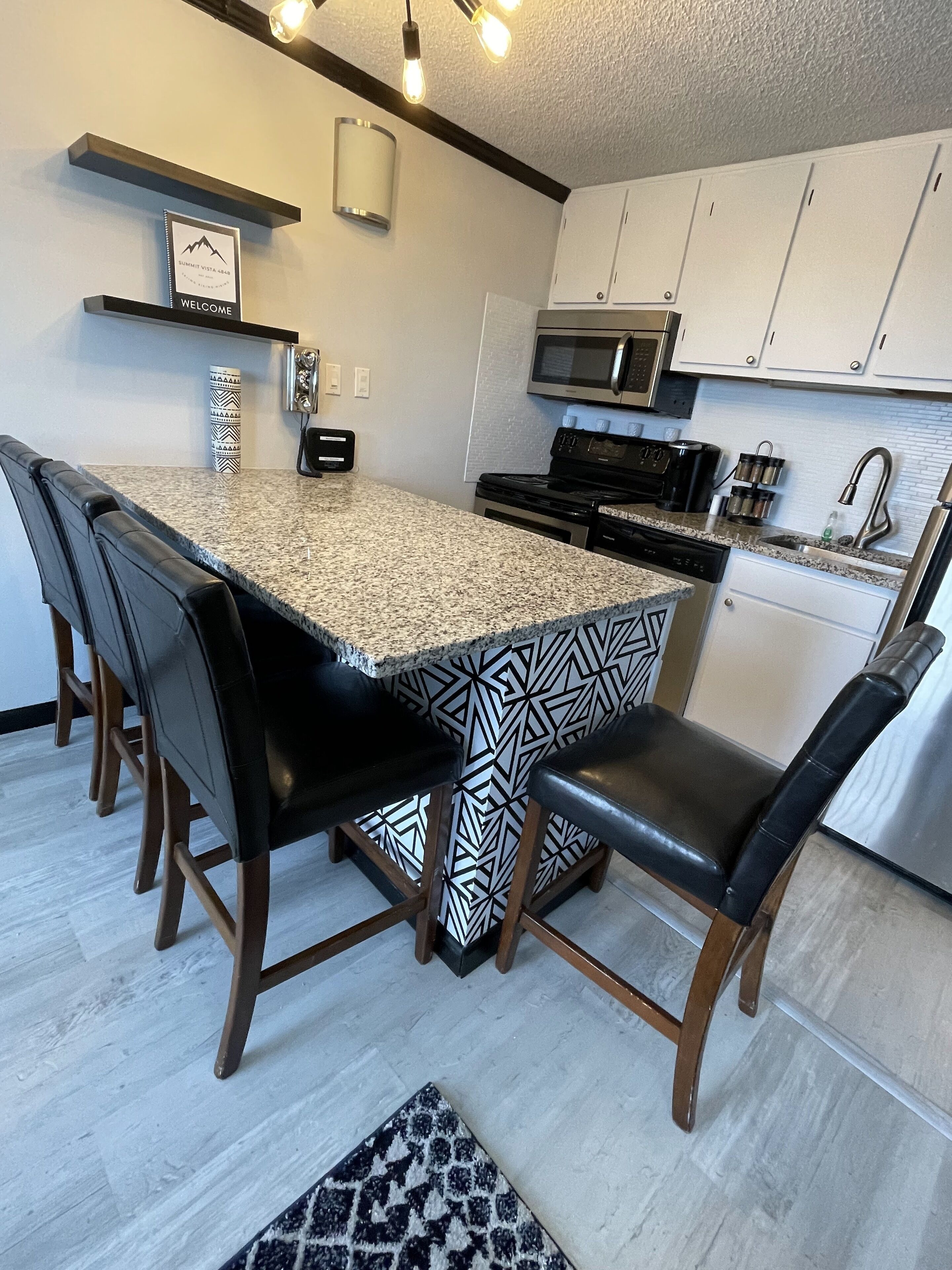 A modern kitchen with a granite countertop, black and white patterned stools, a built-in microwave, and a coffee machine.