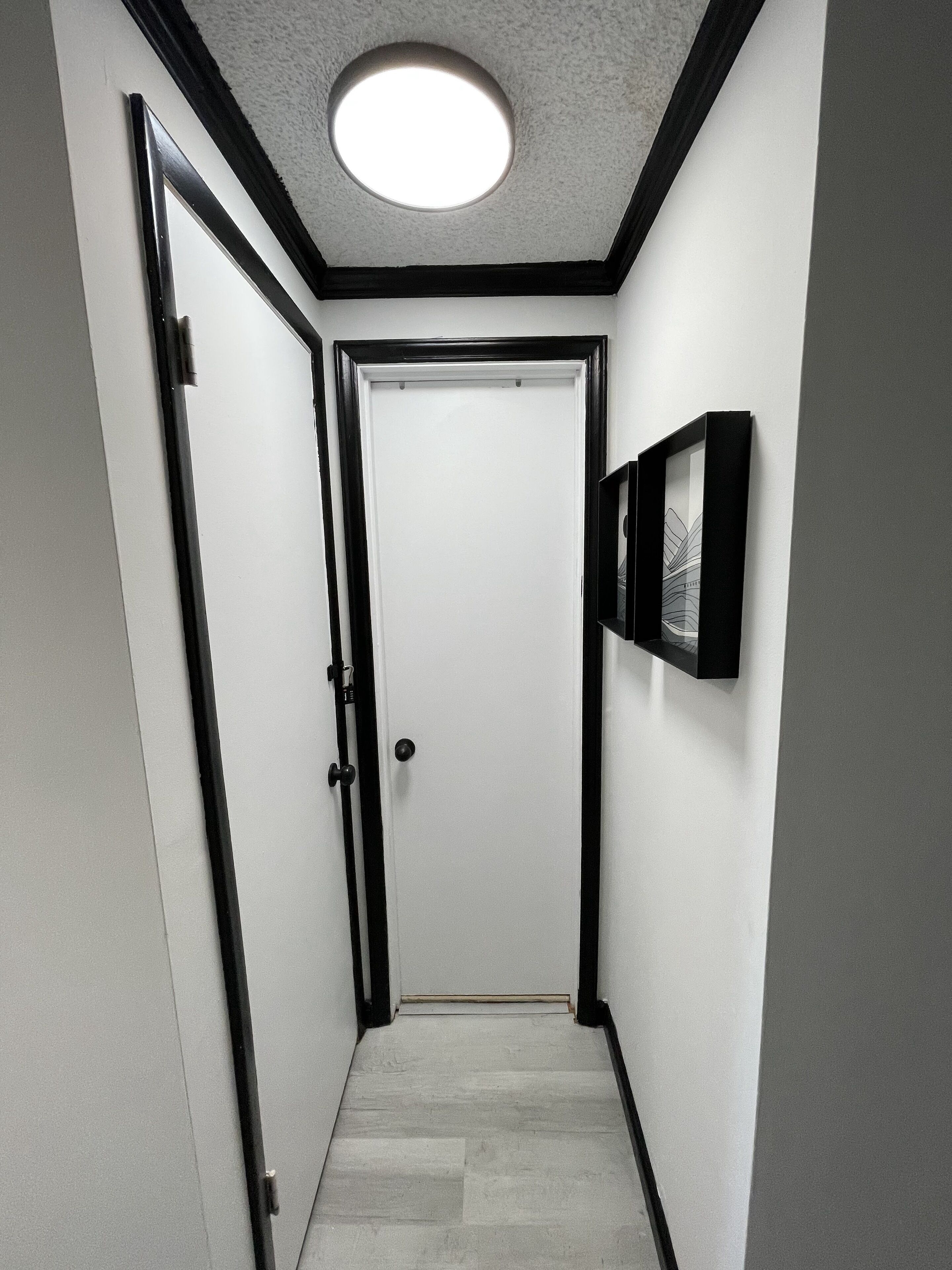 Un corridoio stretto con una porta bianca in fondo, un quadro con cornice nera sulla parete e una luce a soffitto.
