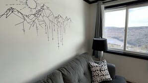 Une pièce avec un canapé gris, un coussin à motifs, une décoration murale représentant des montagnes et un soleil, et une fenêtre donnant sur une forêt et un plan d’eau.