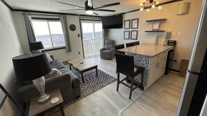 Un appartement moderne comprenant un salon, un coin repas et une cuisine. Il est doté d’un ventilateur de plafond, de grandes fenêtres et offre une vue sur la ville.