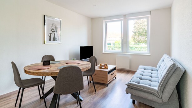 A modern living room with a wooden dining table, grey chairs, a grey sofa, and a television.