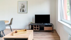 A modern living room with a flat-screen TV, a framed picture on the wall, a wooden coffee table, and a dining area with a gray chair.