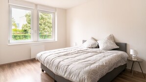 Une chambre à coucher avec un lit, deux fenêtres, une table de chevet avec une lampe, et une vue sur la verdure à l’extérieur.
