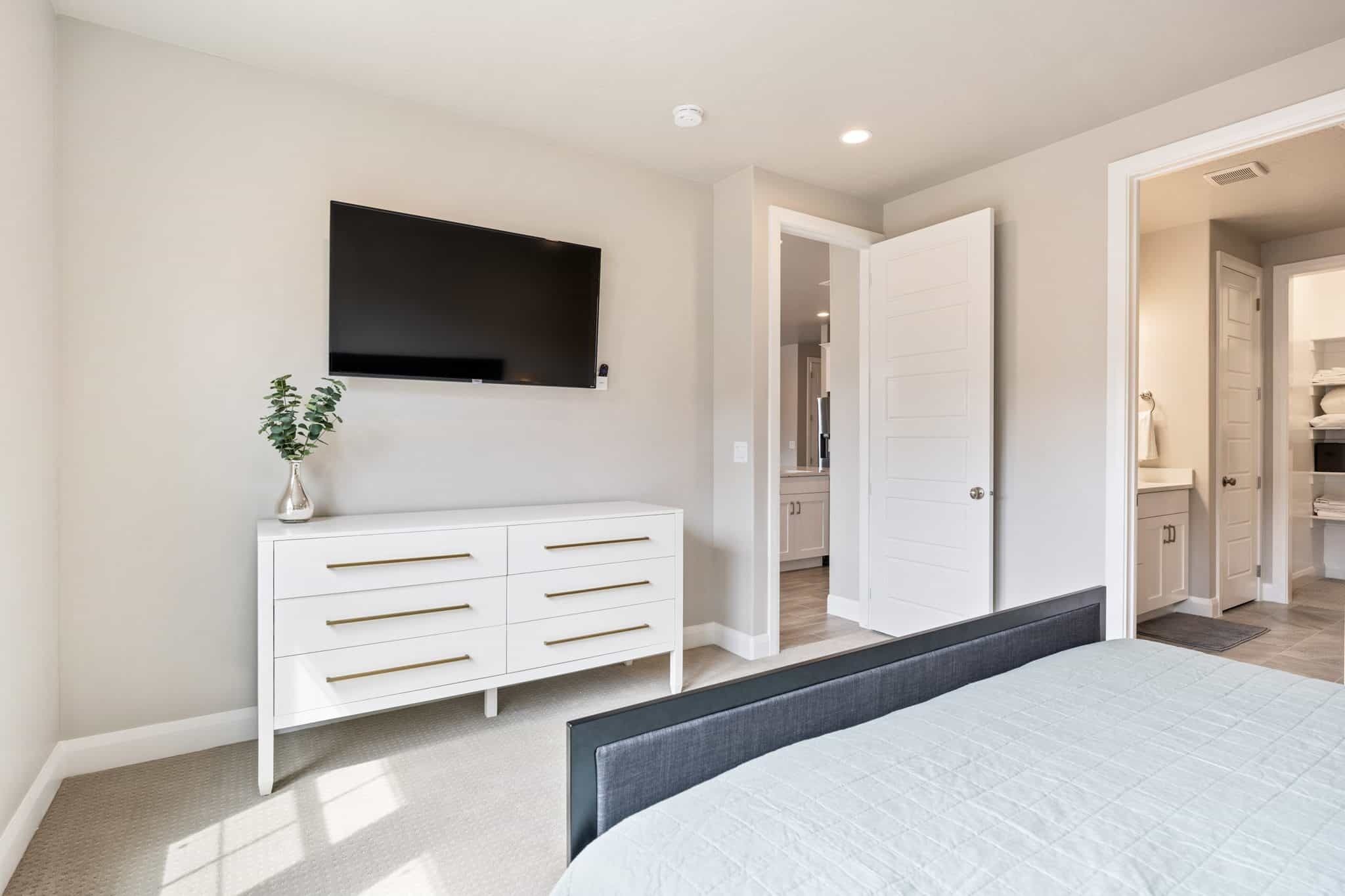 Un dormitorio con cama, cómoda, televisor montado en la pared y puerta que da a un baño.