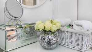 Et badeværelsesbord med et spejl af glas, en dekorativ vase med hvide blomster og en fletkurv med håndklæder.