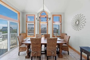 A dining area with a large table and wicker chairs, large windows with wooden frames, and a decorative wall piece. - Oceanview Home with Private Pool, Hot Tub, Game Room, and Nearby Beach Access (Avon)
