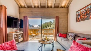 A modern living room with a sofa, coffee table, and a TV. There is a balcony with a view of mountains.