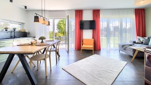 Un salon moderne comprenant un canapé, une table à manger avec des chaises, un téléviseur fixé au mur et une grande fenêtre avec des rideaux.