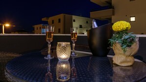 Een tafel met twee glazen amberkleurige vloeistof, een kaars en een fles champagne, voor een gebouw 's nachts.