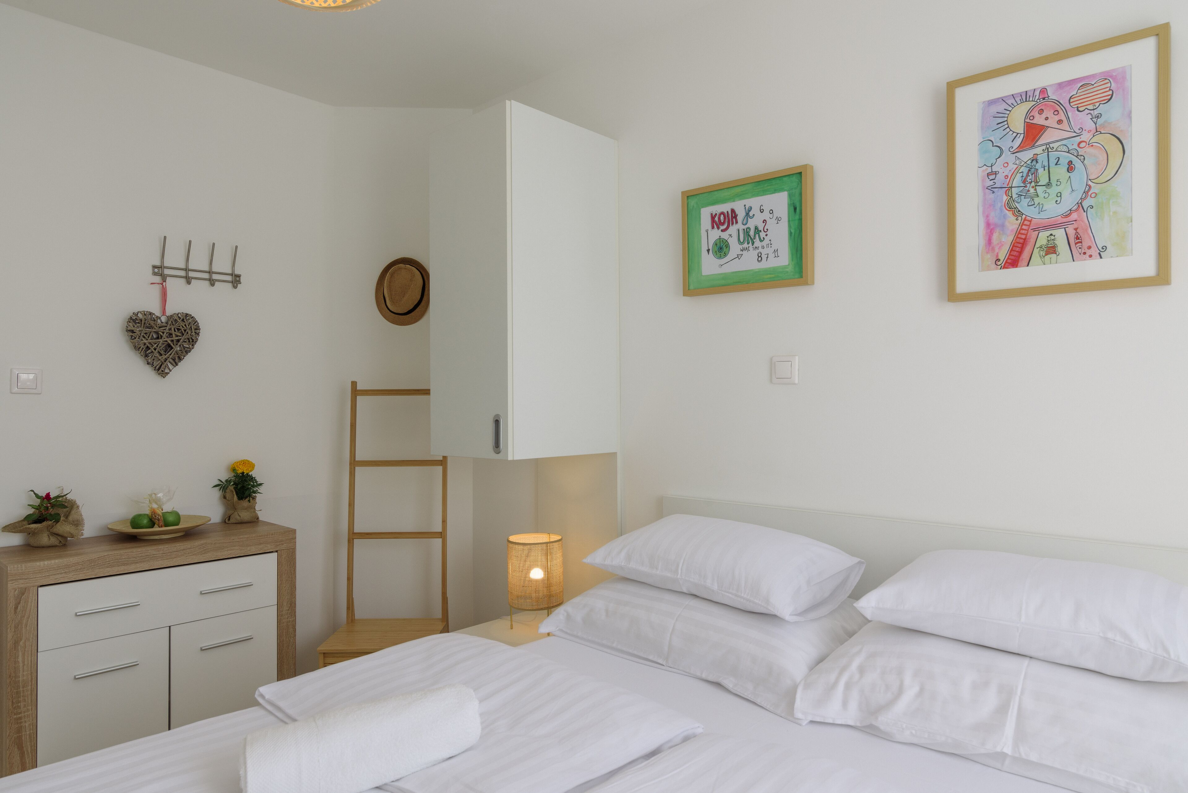 A bedroom with a bed, bedside table, lamp, and wall decorations including a heart-shaped item and framed artwork.