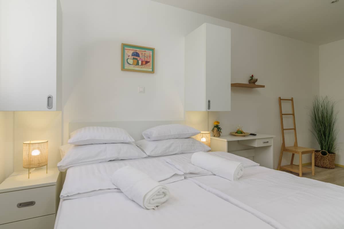 Una cama bien hecha con sábanas blancas, una lámpara de noche, una mesita con una planta y una silla de madera.