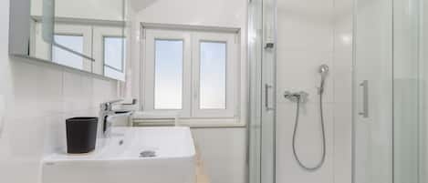 Une salle de bain moderne avec un lavabo blanc, une tasse noire, une cabine de douche en verre et une fenêtre qui laisse entrer la lumière naturelle.