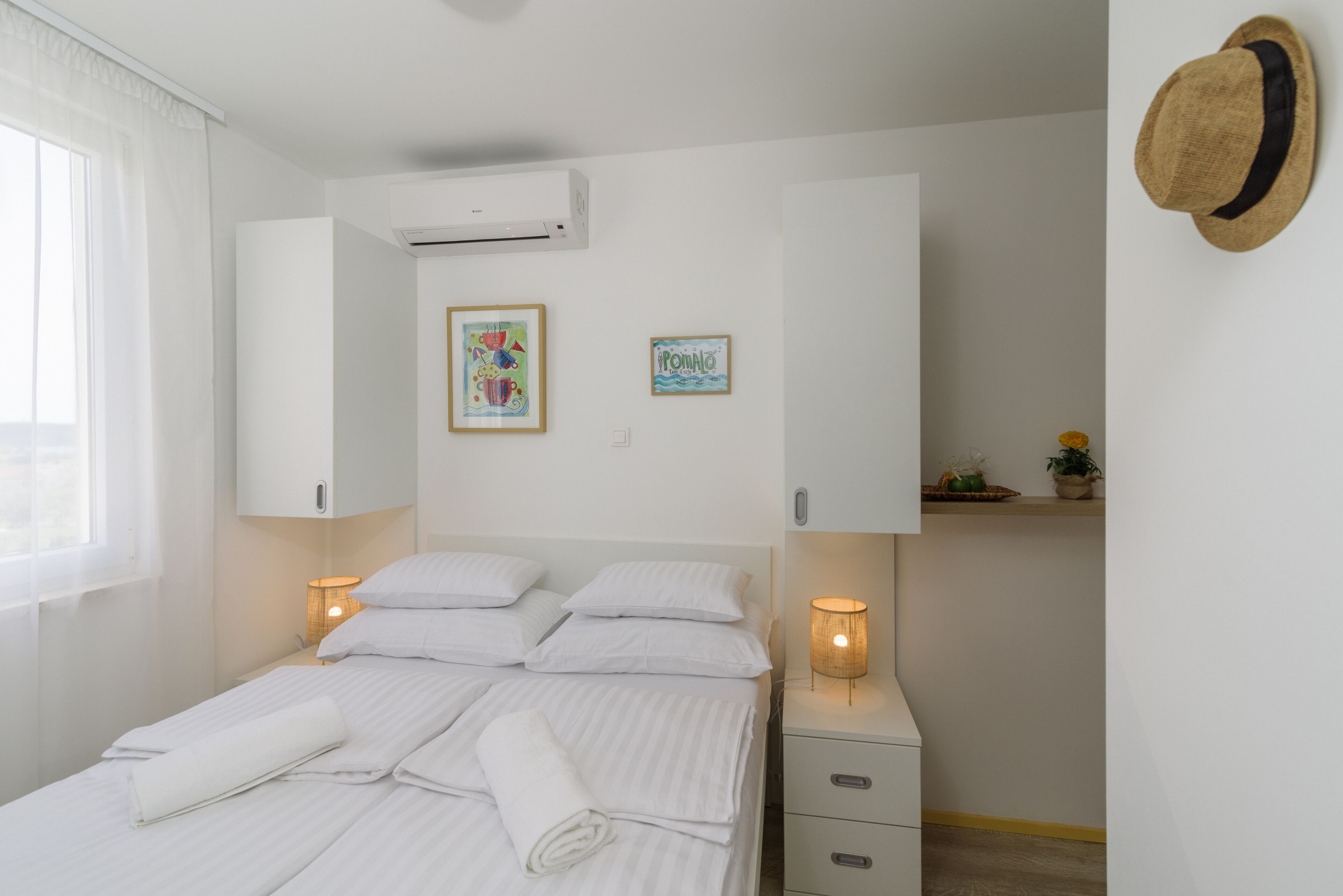 A neatly made bed with white linens, a bedside lamp, a small bedside table with a lamp, and a wall-mounted hat.