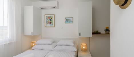 A neatly made bed with white linens, a bedside lamp, a small bedside table with a lamp, and a wall-mounted hat.
