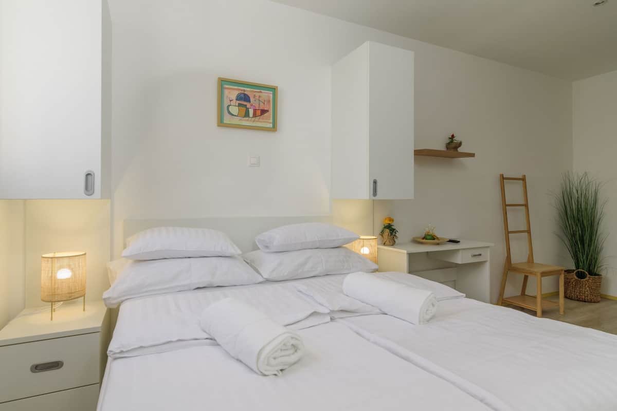Una cama bien hecha con sábanas blancas, una lámpara de noche, un estante con una planta y un cuadro en la pared.