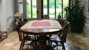 A dining area with a large wooden table, six chairs, and a patterned table runner. There is a stone wall and a large window with a view of greenery.