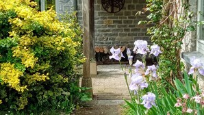 A stone house with a tiled roof, a covered porch, and a garden with yellow and purple flowers.
