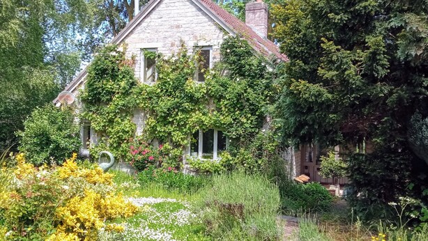 A stone house with a steep roof, surrounded by lush greenery and flowering plants.