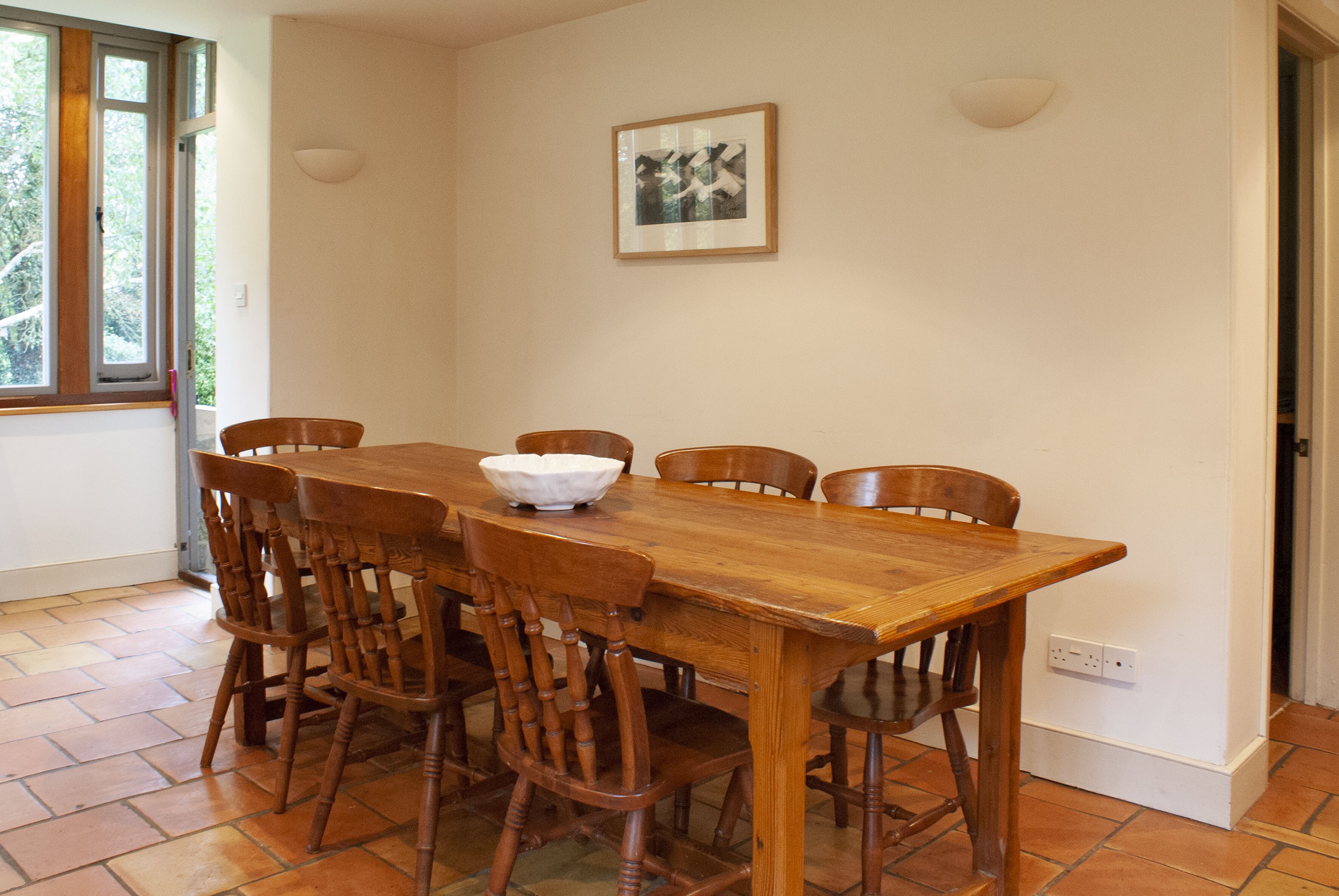 A dining area with a wooden table and chairs, a white bowl on the table, and a framed picture on the wall.