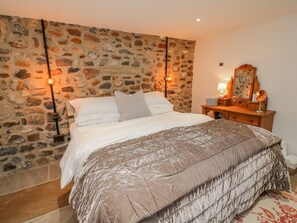 Un dormitorio con pared de piedra, una cama con colcha color beige, un tocador de madera con espejo y dos lámparas de pared.
