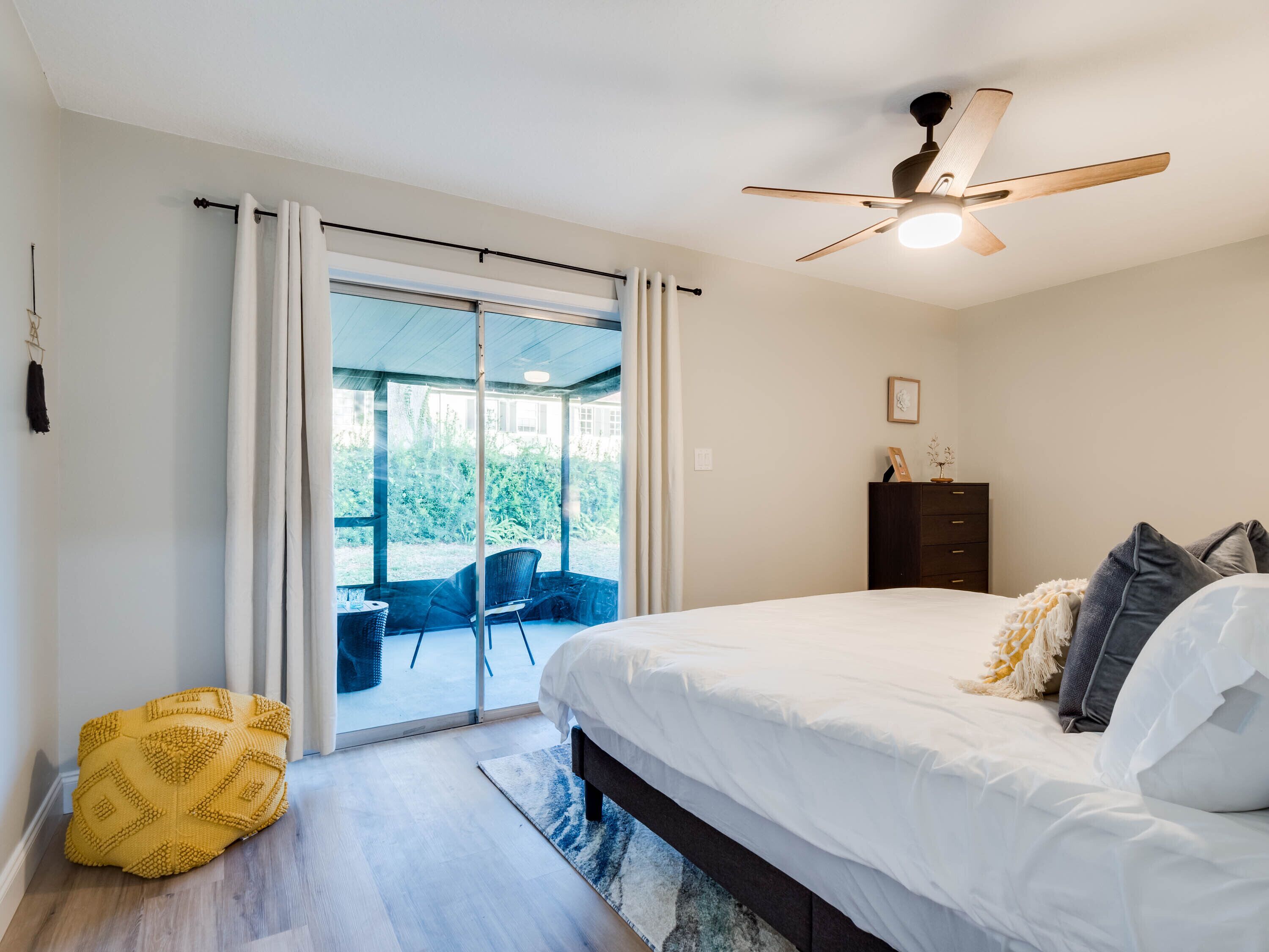 Ein Schlafzimmer mit einem Bett, einem Deckenventilator, einer dunkelholznen Kommode und Blick durch eine Glastür auf einen Außenbereich mit einem Stuhl.