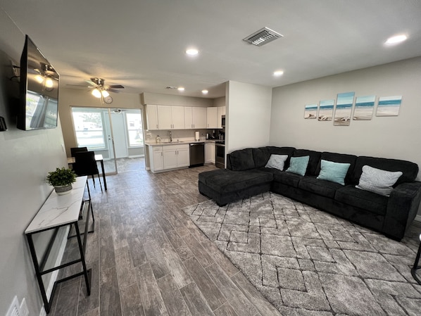 A modern living room with a sectional sofa, a dining area with a table and chairs, and a kitchen in the background.