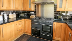 A kitchen with wooden cabinets, a stainless steel range hood, a gas stove, and a microwave. - 11 Bro Celyn (Holyhead)