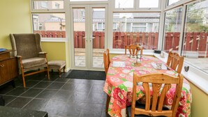 A sunroom with a table and chairs, a wooden chair, a cabinet, and a view of a red fence through large windows. - 11 Bro Celyn (Holyhead)