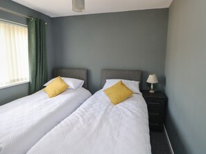 A hotel room with two single beds, a nightstand with a lamp, and a window with curtains. - 11 Bro Celyn (Holyhead)