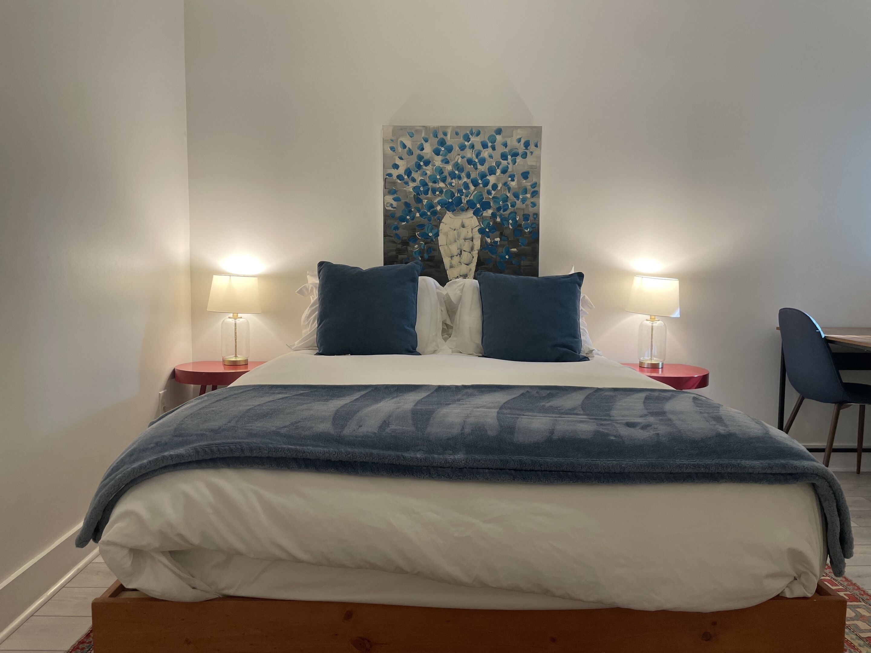 Una cama bien hecha con una colcha azul, dos lámparas de noche, una obra de arte en la pared y una silla.