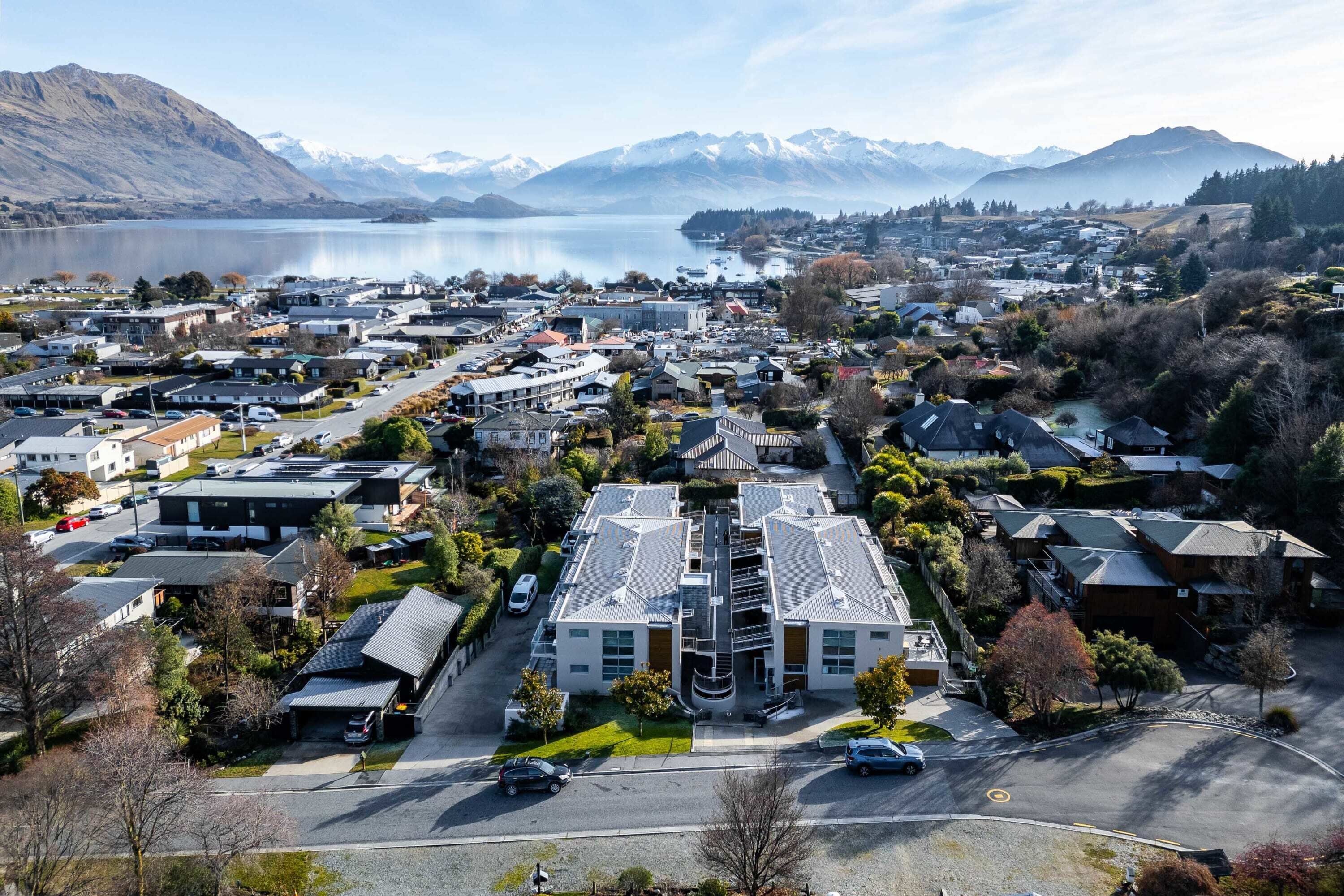 An aerial view of a residential area with houses, a central building with a distinctive roof, and a lake with mountains in the background.