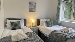 Et dobbeltværelse med to senge, et natbord med lampe og et vindue med gardiner.